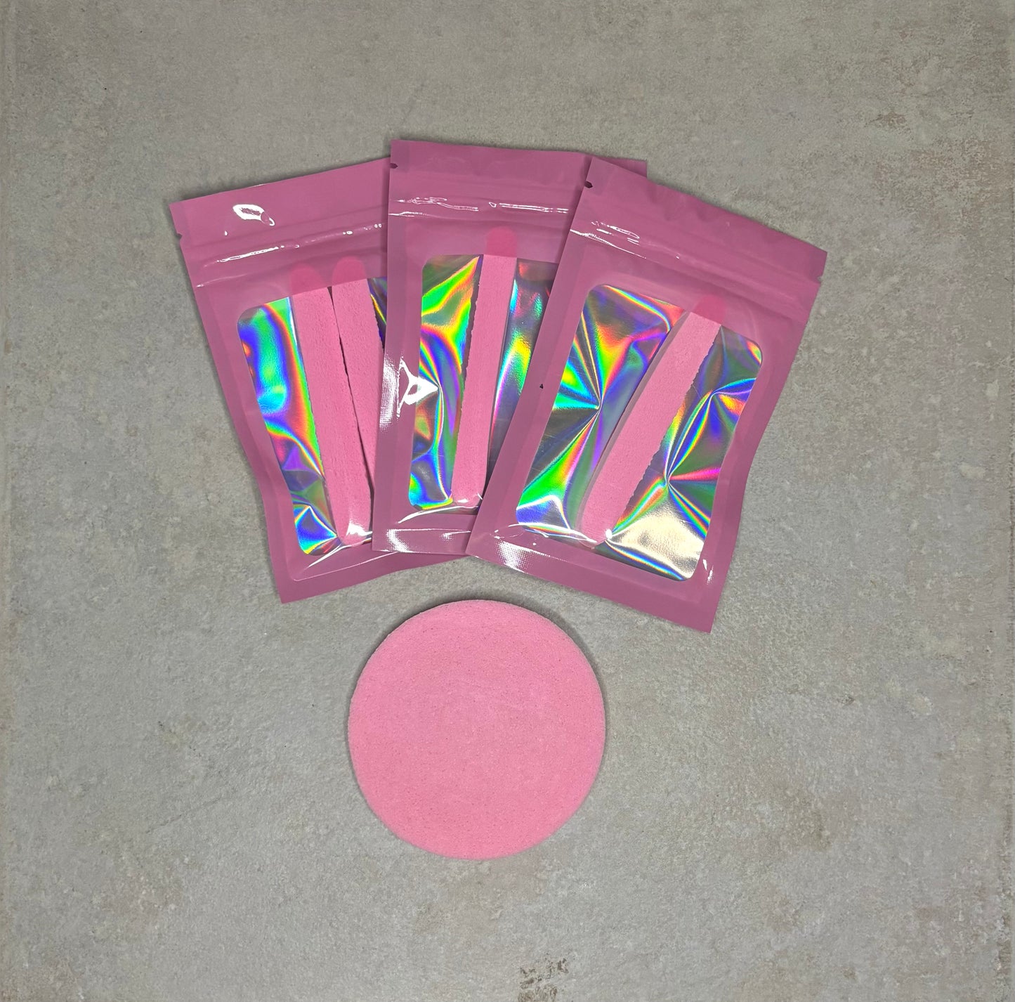 Three pink holographic packaging pouches displayed on a textured surface, each containing pink makeup applicator sponges. A round pink sponge sits below the pouches, showcasing the beauty application accessories set.