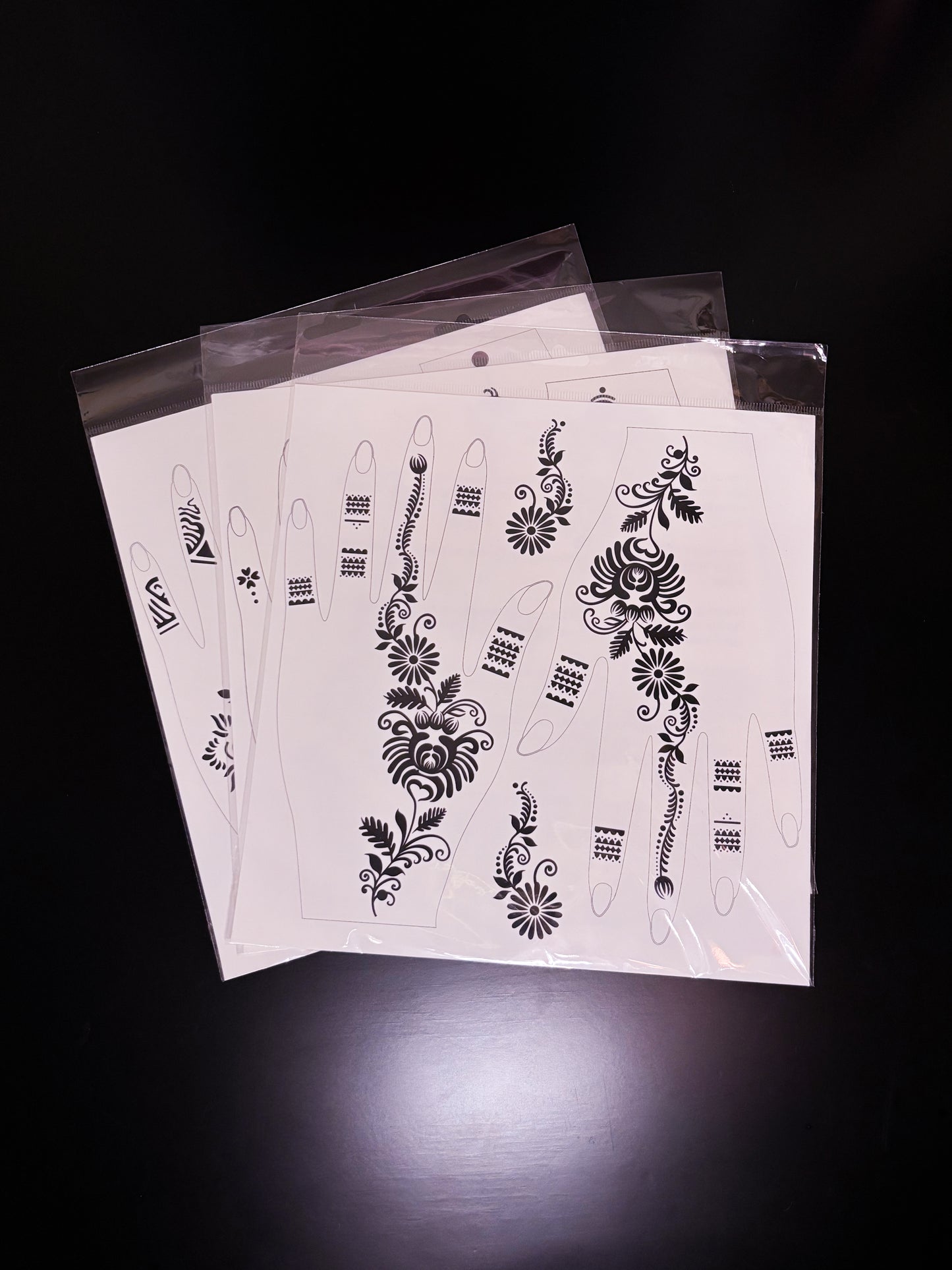 A fanned-out stack of temporary tattoo or henna stencil sheets packaged in clear plastic sleeves, displaying black floral and geometric patterns designed to fit the shape of a hand and fingers.