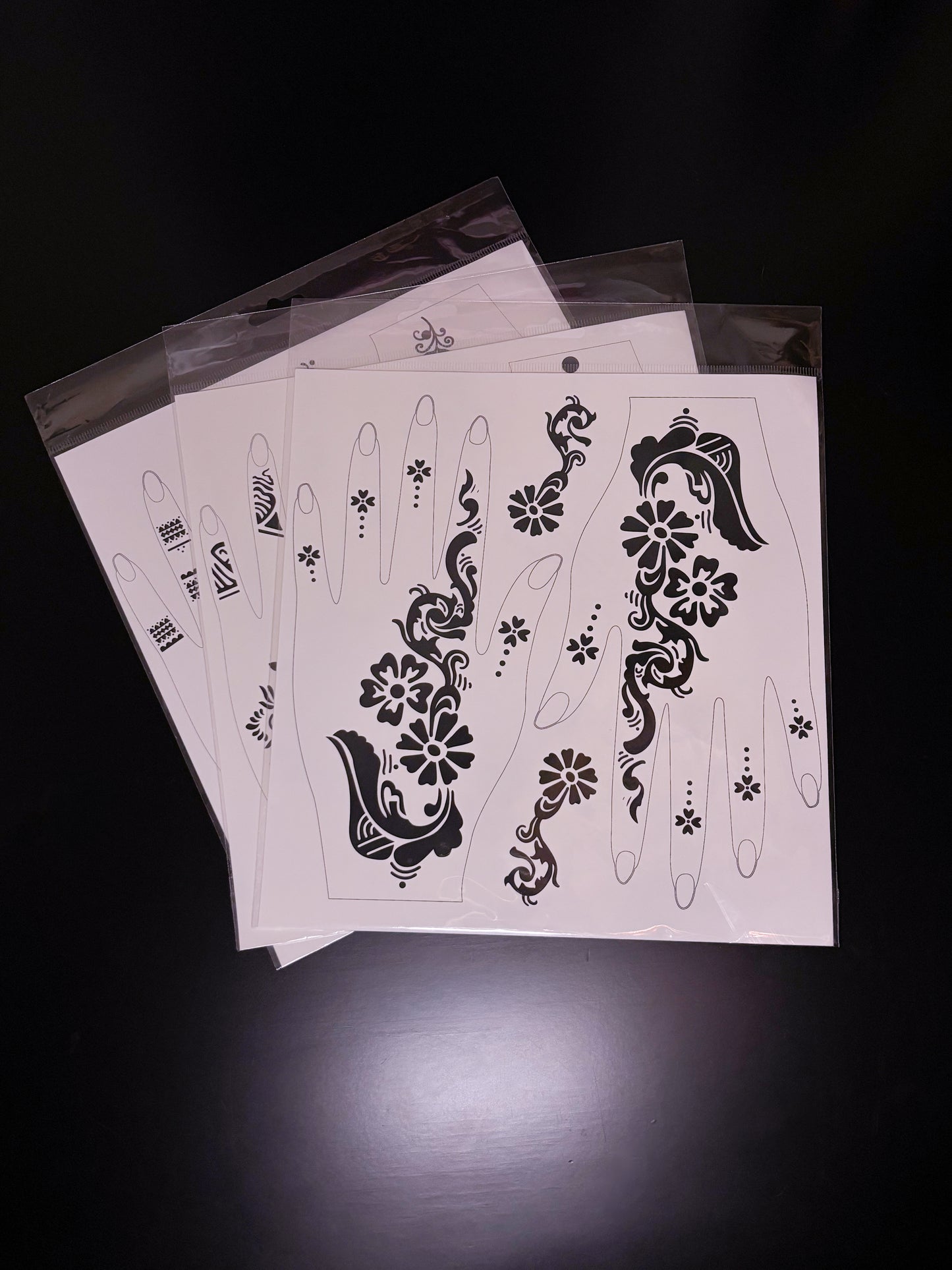 Alt Text

A fanned-out stack of temporary tattoo or henna stencil sheets in clear cellophane packaging, displayed on a dark surface. The top sheet shows bold, black floral patterns with swirling vine details and delicate stippled dots designed to fit the contours of a left and right hand.