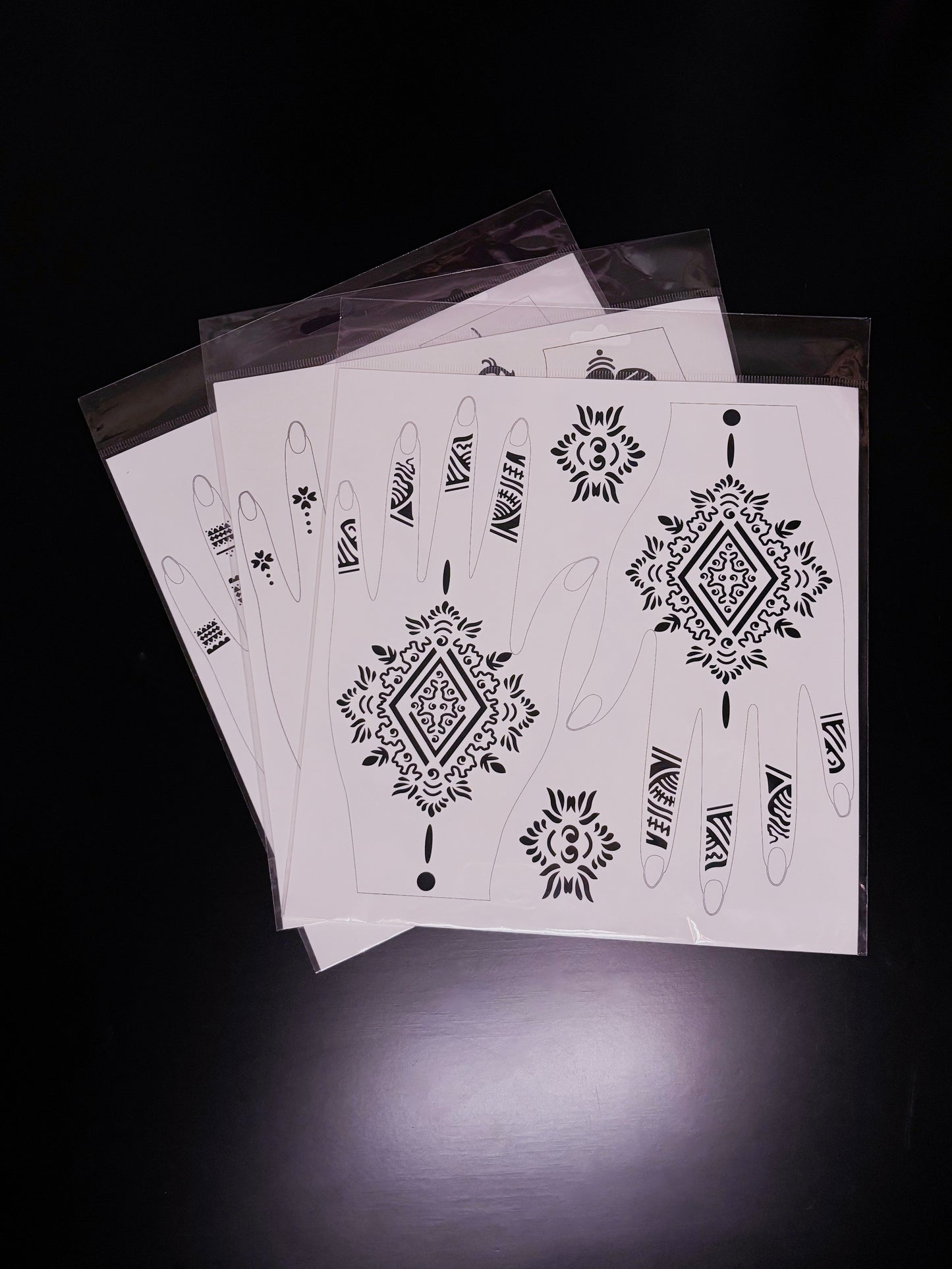A fanned-out stack of temporary tattoo or henna stencil sheets in clear cellophane sleeves, displayed on a dark reflective surface. The top sheet features a geometric and tribal-inspired design, highlighted by a large central diamond mandala for the back of the hand and matching linear, abstract patterns for the fingers.
