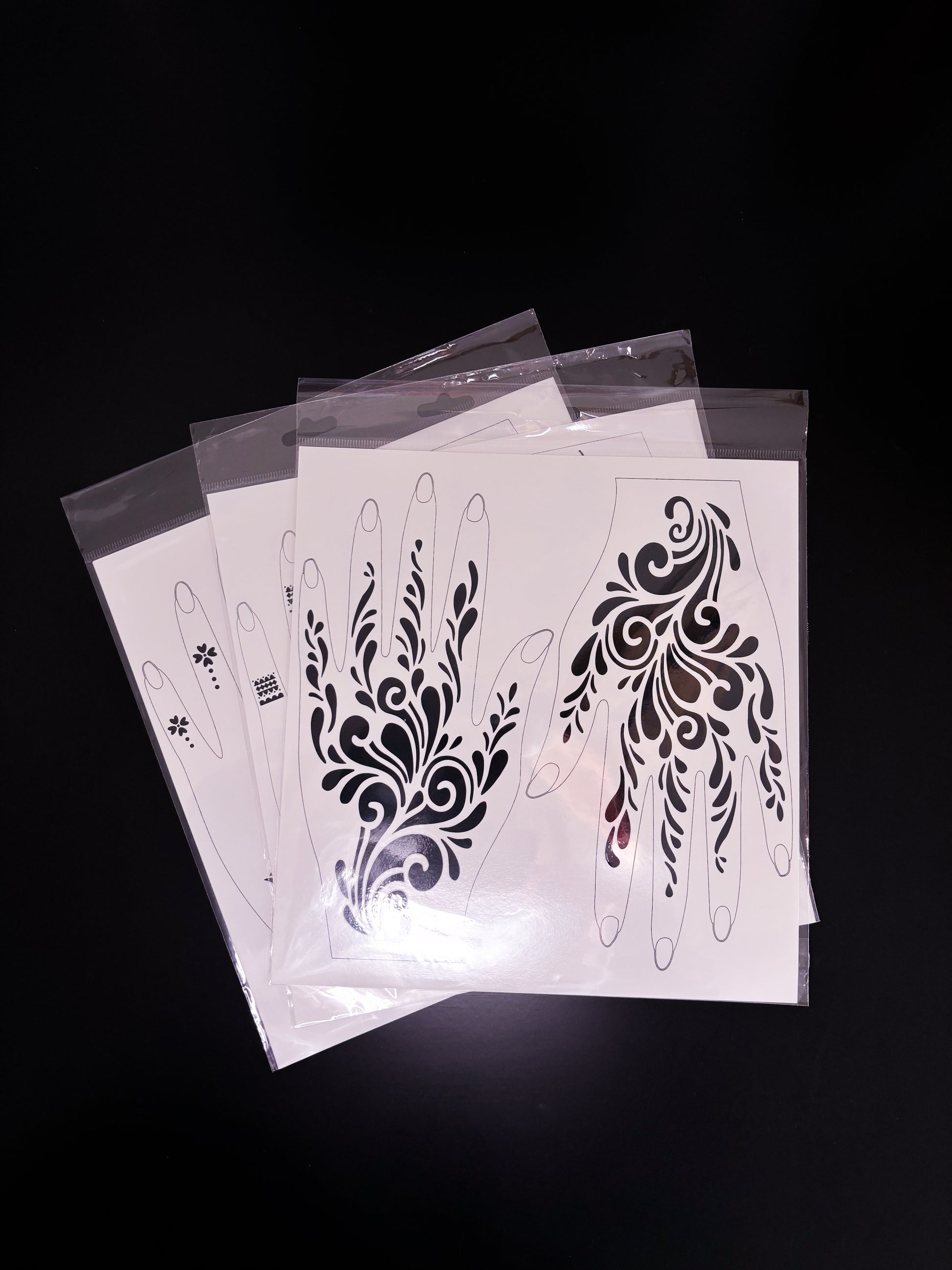A fanned-out stack of temporary tattoo or henna stencil sheets in clear cellophane sleeves, resting on a dark reflective surface. The top sheet displays an abstract, flowing design featuring elegant curls, swirling waves, and teardrop flourishes that wind upward across the back of the hands and fingers.