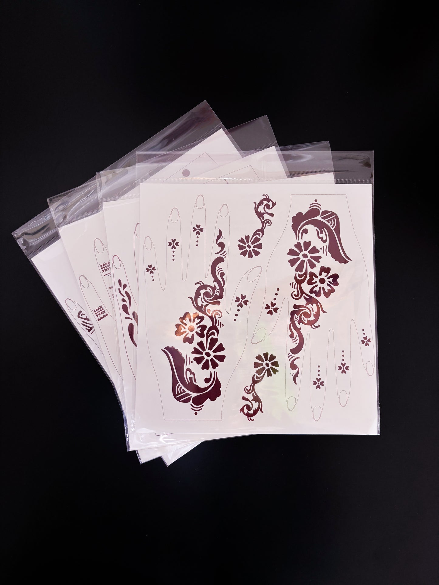 A fanned-out stack of temporary tattoo or henna stencil sheets in clear cellophane packaging, resting on a dark surface. The top sheet displays a bold floral trail layout with large daisy-like blossoms, sweeping paisley curves near the wrist, and delicate dotted flower accents for the fingers.