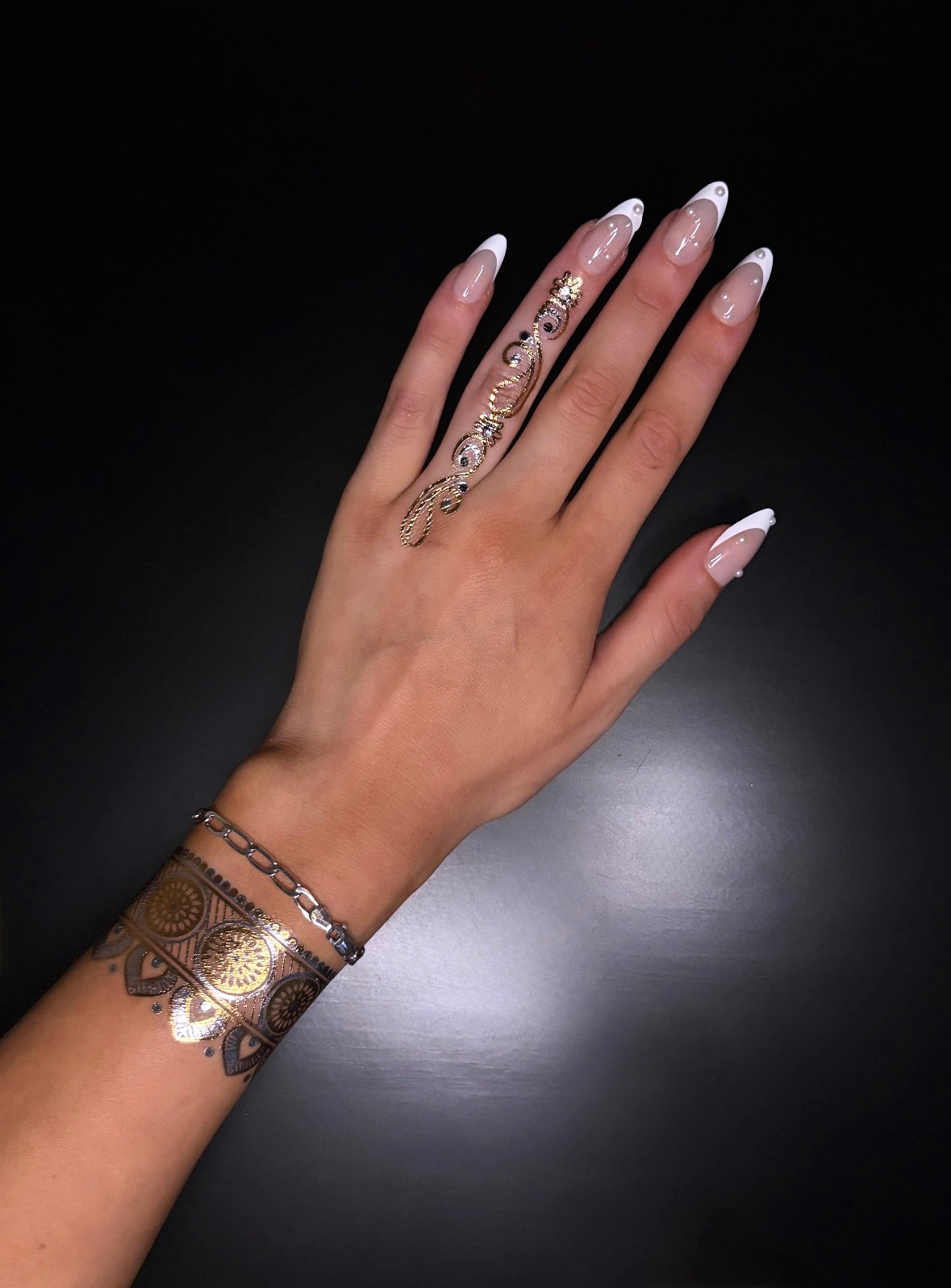 A top-down view of a hand with a manicured French tip nail design, a silver chain link bracelet, and an ornate, metallic gold and black cuff-style tattoo on the wrist. One finger features an intricate, full-length silver ring.