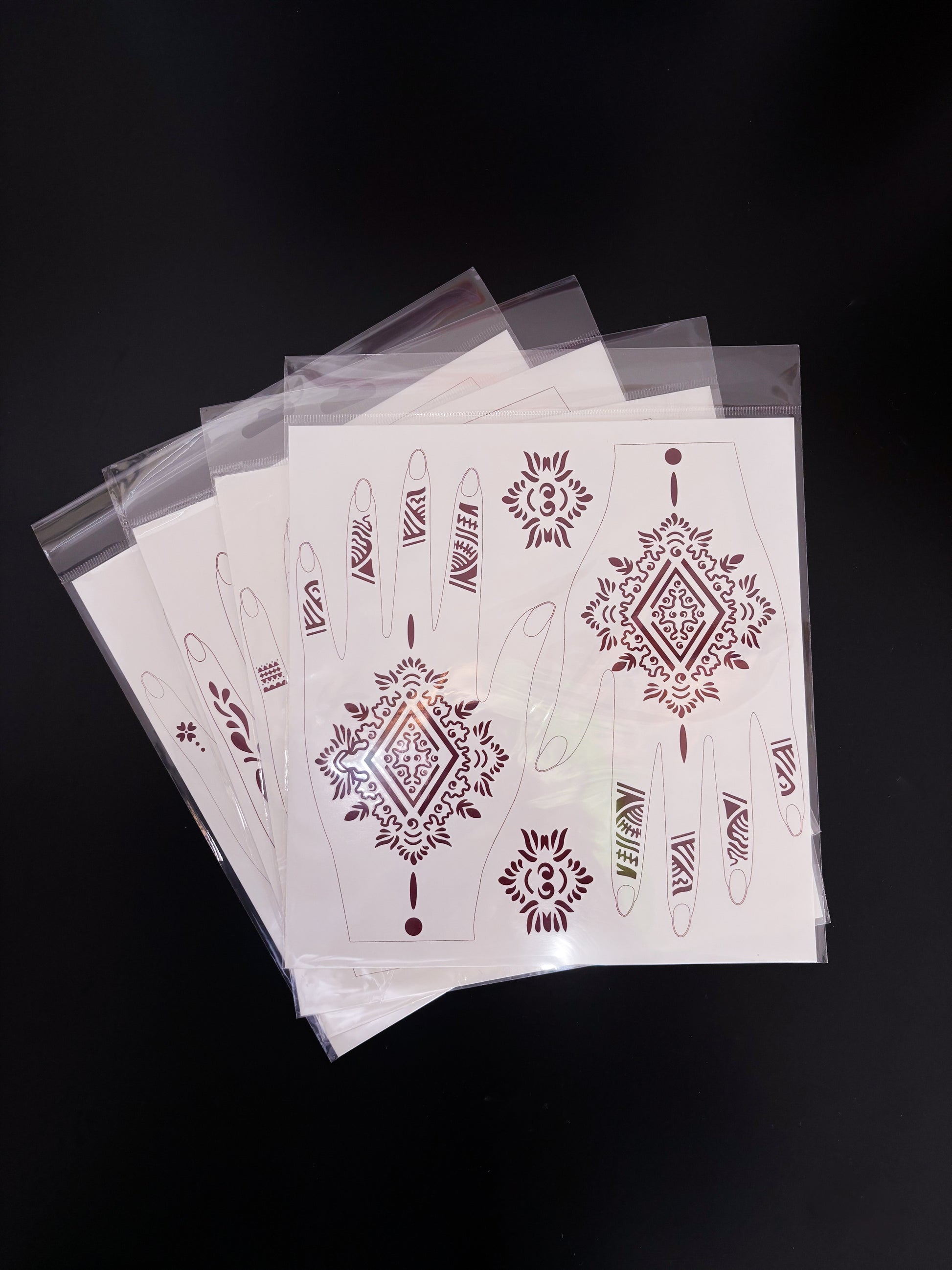 A fanned-out stack of temporary tattoo or henna stencil sheets in clear cellophane sleeves, laid on a dark surface. The top sheet features a sharp geometric design in a rich, dark maroon or burgundy ink, highlighting a large central diamond mandala for the back of the hands, abstract tribal-style finger bands, and two independent accent medallions.