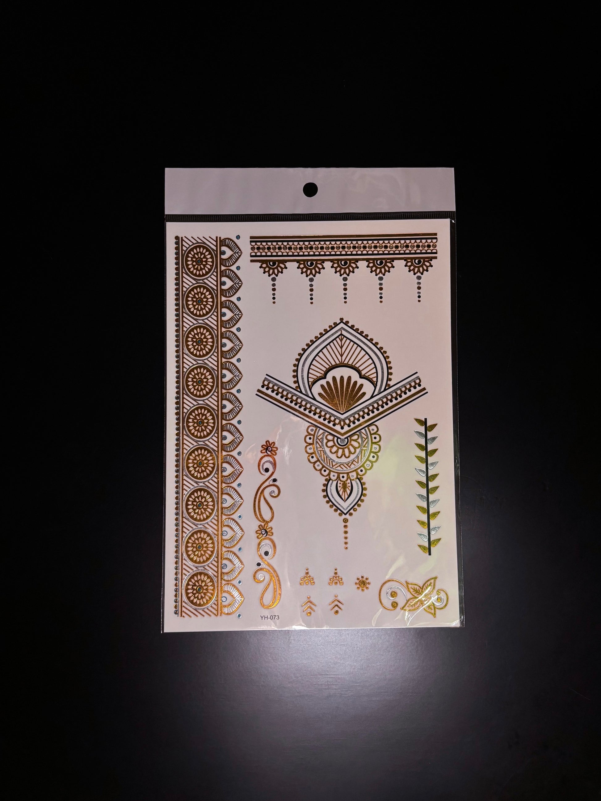 A single metallic gold temporary henna tattoo sheet displayed against a black background on a computer screen. The sheet features intricate decorative designs including floral borders, geometric patterns, paisley-inspired elements, and small accent details in reflective gold and brown tones.