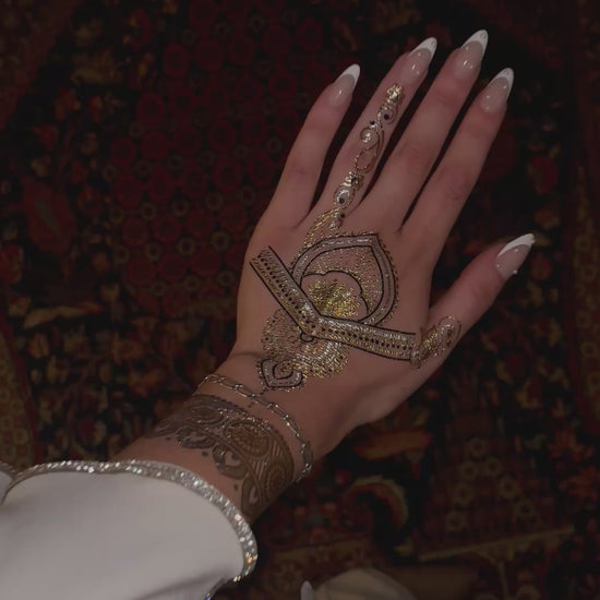 Close-up video of a hand wearing a metallic gold temporary henna tattoo under flash lighting. The intricate design sparkles and reflects light, highlighting detailed floral and geometric patterns with a glossy metallic finish.