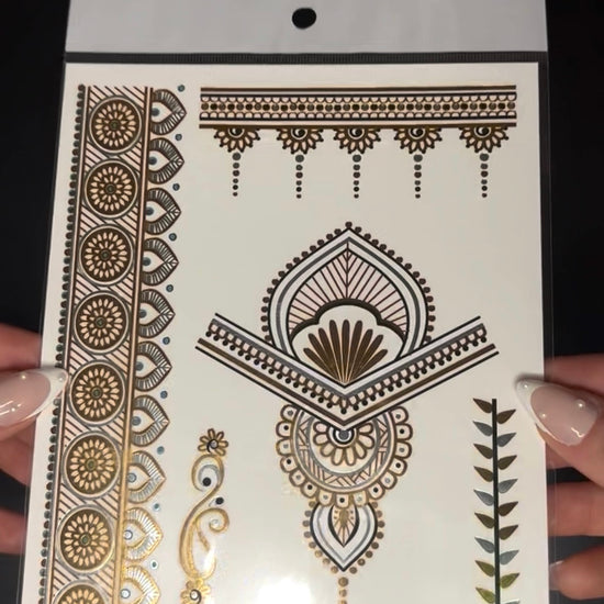 Close-up video of a metallic gold temporary henna tattoo sheet being held and tilted in front of the camera, revealing reflective gold details and intricate floral and geometric patterns that shimmer under the light.