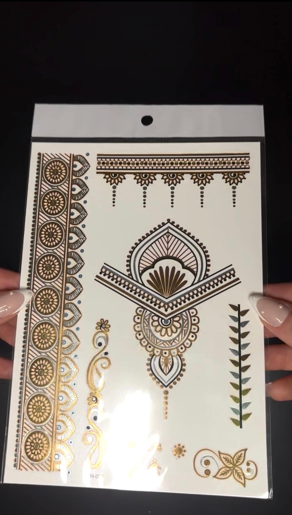 Close-up video of a metallic gold temporary henna tattoo sheet being held and tilted in front of the camera, revealing reflective gold details and intricate floral and geometric patterns that shimmer under the light.